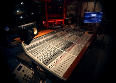 Close-up overhead view of a large professional mixing console with rows of faders, knobs and channel strips, flanked by studio monitor speakers and a computer screen displaying a DAW desktop.