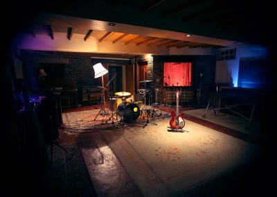 Atmospheric recording studio live room with a miced-up drum kit on a patterned rug, a sunburst electric guitar on a stand, a floor lamp, exposed wooden ceiling beams, and coloured ambient lighting in purple and blue tones.