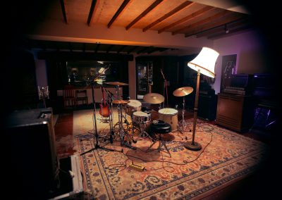 Recording studio live room showing a drum kit surrounded by microphone stands and cables on a decorative rug, with a floor lamp providing warm light, a guitar leaning against a mic stand, and vintage amplifiers visible to the right.
